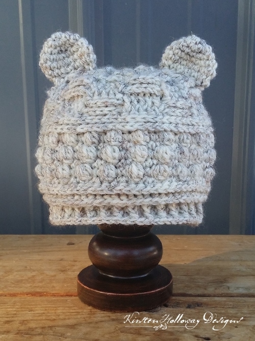 Cream color crochet beanie with bear ears displayed on wooden hat display with dark gray background.