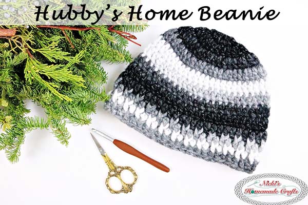 A black, gray and white crochet beanie next to pine needle branch, yarn scissors and crochet hook displayed on white background 