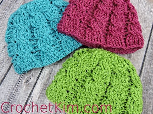 Three cable crochet newborn beanies in colors blue, pink and green on wood background