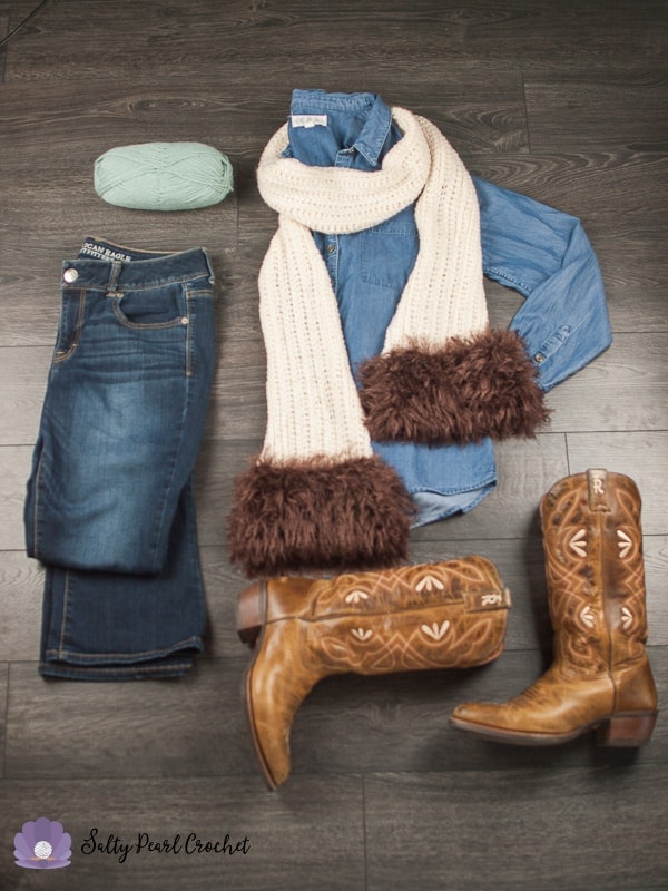 Flat lay of skein of light blue yarn, folded jeans, a jean shirt folded with cream knit scarf with brown faux fur muff wrapped around jean shirt and western cowboy boots on wooden floor. 