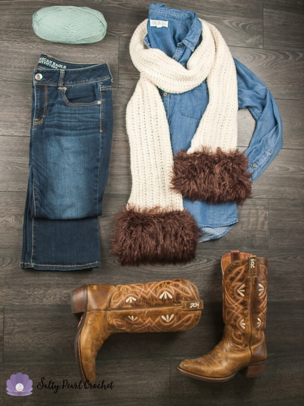Flat lay of skein of light blue yarn, folded jeans, a jean shirt folded with cream knit scarf with brown faux fur muff wrapped around jean shirt and western cowboy boots on wooden floor. 