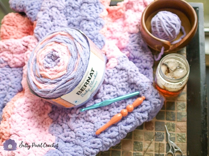 Close up of pink and purple crochet blanket on a table with a skein of pink and purple yarn, a metal crochet hook, and a wooden crochet hook on top of the blanket. 