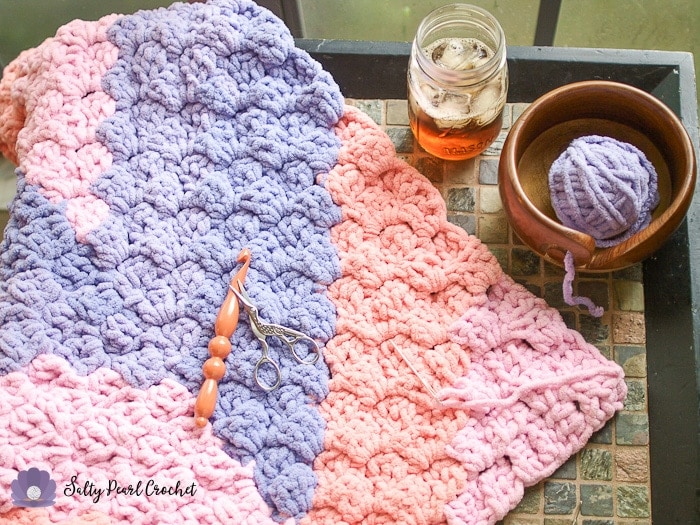 Pink and purple crochet blanket on a table with a wooden crochet hook and scissors on the blanket. A glass of iced tea and yarn bowl with purple yarn in it are also on the table next to the blanket. 