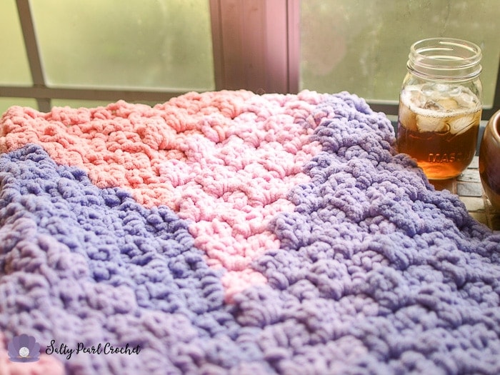 Close up of pink and purple crochet blanket on a table with a glass of iced tea. 