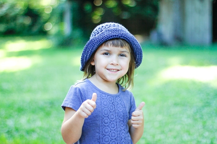 Little girl outside wearing a purple crochet bucket hat giving two thumbs up.