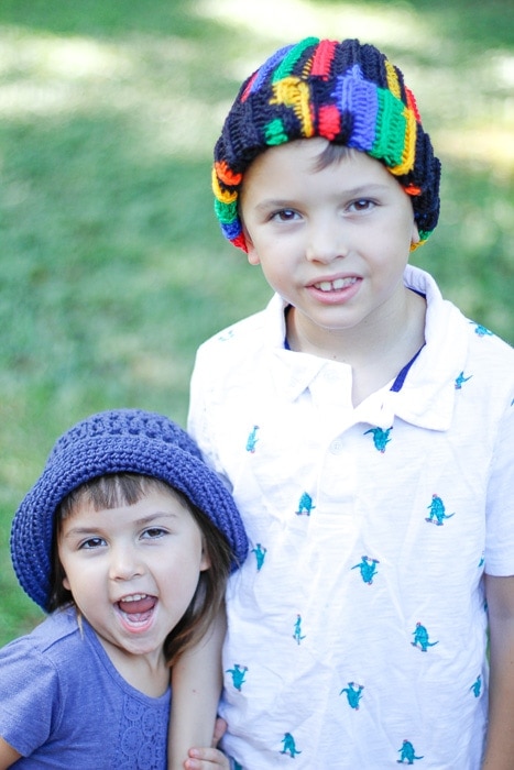 Little girl wearing a purple crochet hat standing next to a little boy wearing a multi-colored crochet beanie. 