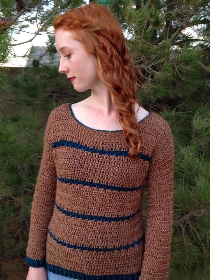 Woman wearing crochet brown sweater with navy stripes standing in front of pine trees. 