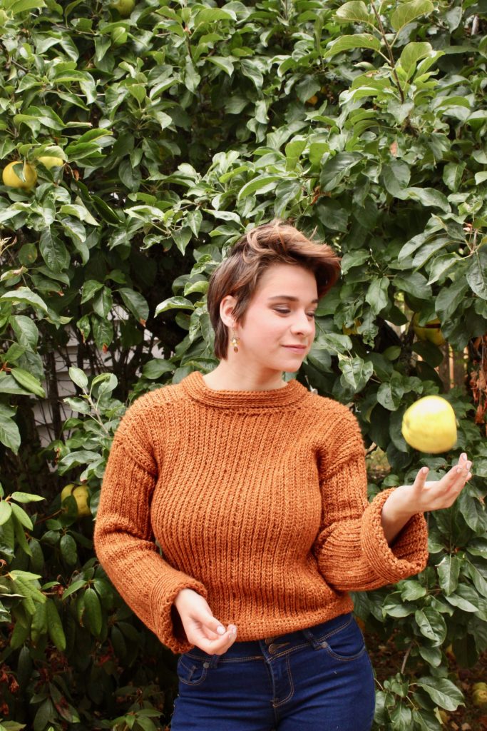 Woman wearing a burnt orange crochet sweater tossing an apple in the air standing in front of large green bush. 