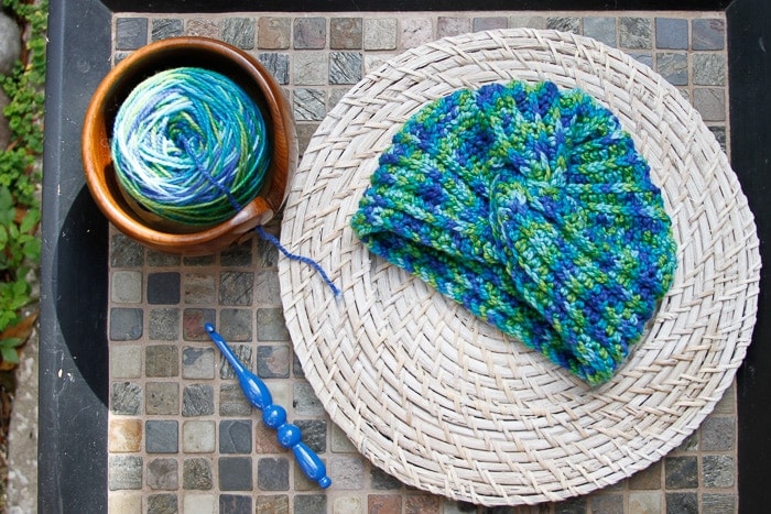 Variegated blue and green crochet turban displayed on wicker with blue crochet hook and blue and green variegated yarn in wooden yarn bowl on tile table. 