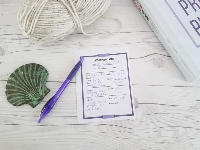 A crochet project note card on a table with a purple pen and a ball of white yarn.