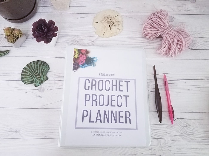 A printable crochet planner on a desk with a crochet hook, a pen, and yarn.