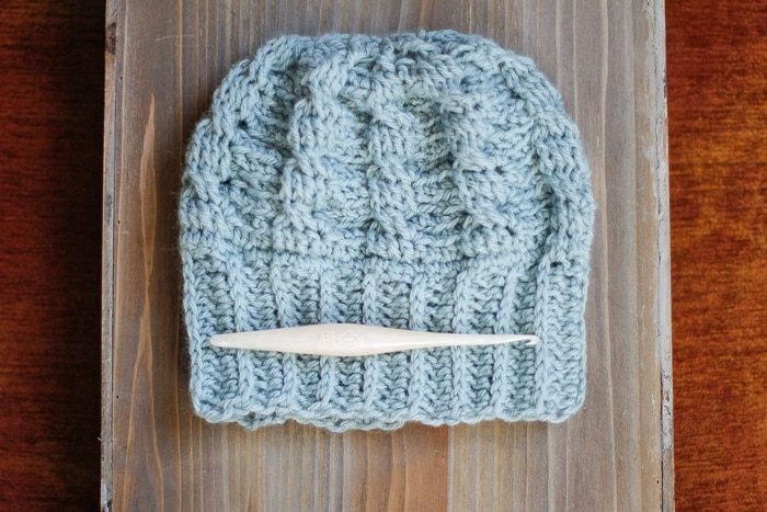 A white crochet hook lying on top of a cabled crochet hat.
