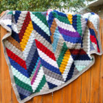 A multicolored herringbone c2c crochet scrap blanket hanging on a wooden fence.
