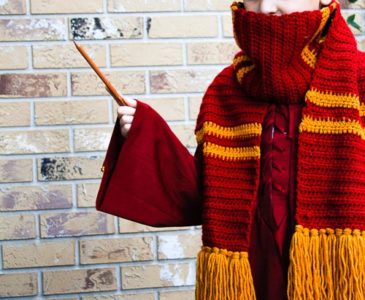 Boy wearing a Gryffindor crochet scarf and waving a wand in front of a brick wall.