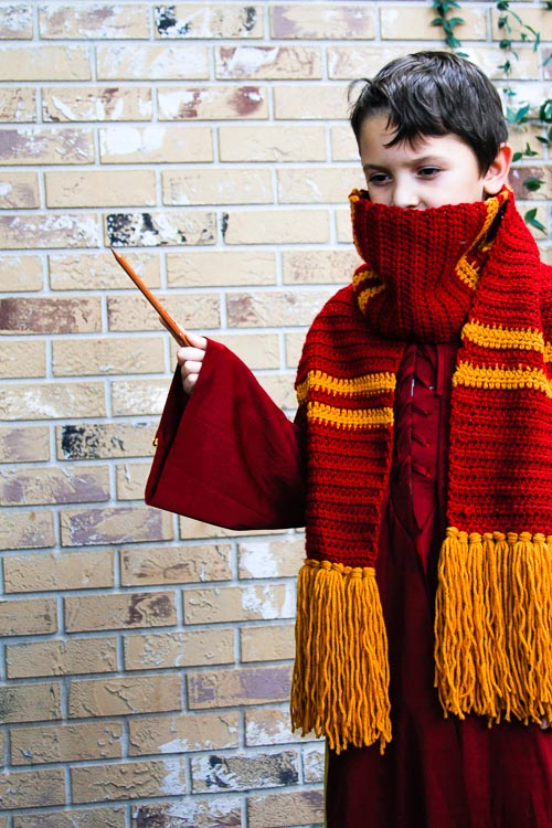 Boy wearing a Gryffindor crochet scarf and waving a wand in front of a brick wall.