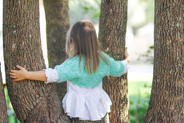 Young girl wearing a green bolero and a white ruffled shirt standing in some trees.