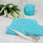 A blue crochet dishcloth with a crochet hook and fancy scissors