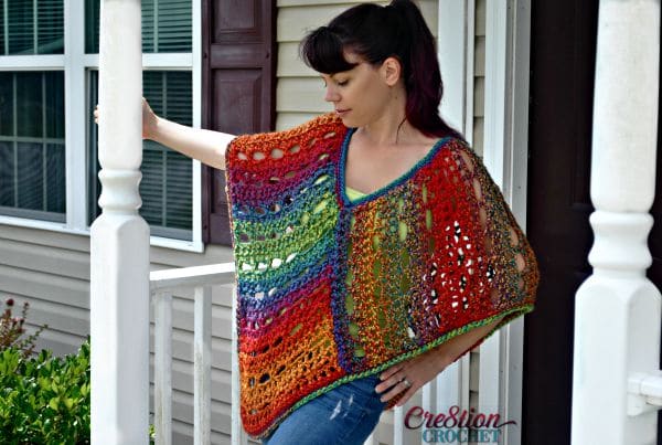 A woman standing on a front porch wearing a mutli-colored crochet poncho. 