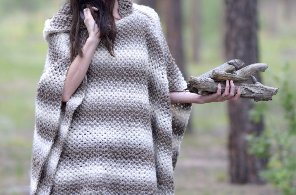 A woman standing outside wearing a neutral color striped crochet poncho holding small log.