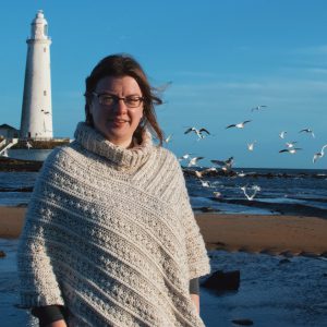 Woman wearing a cream crochet poncho standing in front of a lighthouse on beach. 