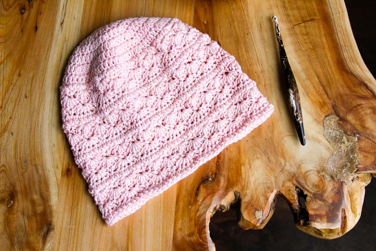 A light pink, highly textured crochet hat on a wood table placed next to a crochet hook. 