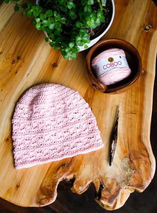 Light pink, highly textured crochet hat on wooden table placed next to a wooden crochet hook, a wooden yarn bowl with pink yarn in it, and a plant. 