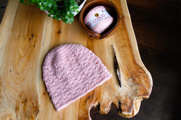 Light pink, highly textured crochet hat on wooden table placed next to a wooden crochet hook, a wooden yarn bowl with pink yarn in it, and a plant. 