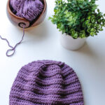 Purple crochet chemo cap beanie laid flat on a white table near yarn and a plant