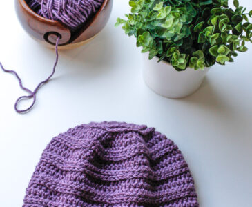 Purple crochet chemo cap beanie laid flat on a white table near yarn and a plant