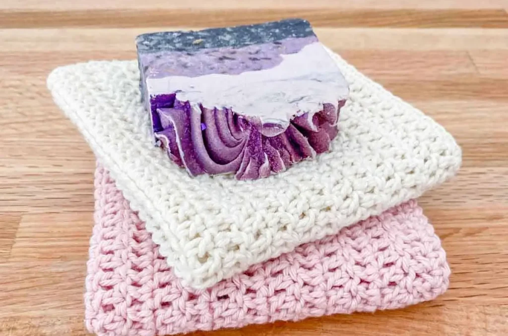 a pink and white crochet washcloth stacked beneath a bar of purple soap