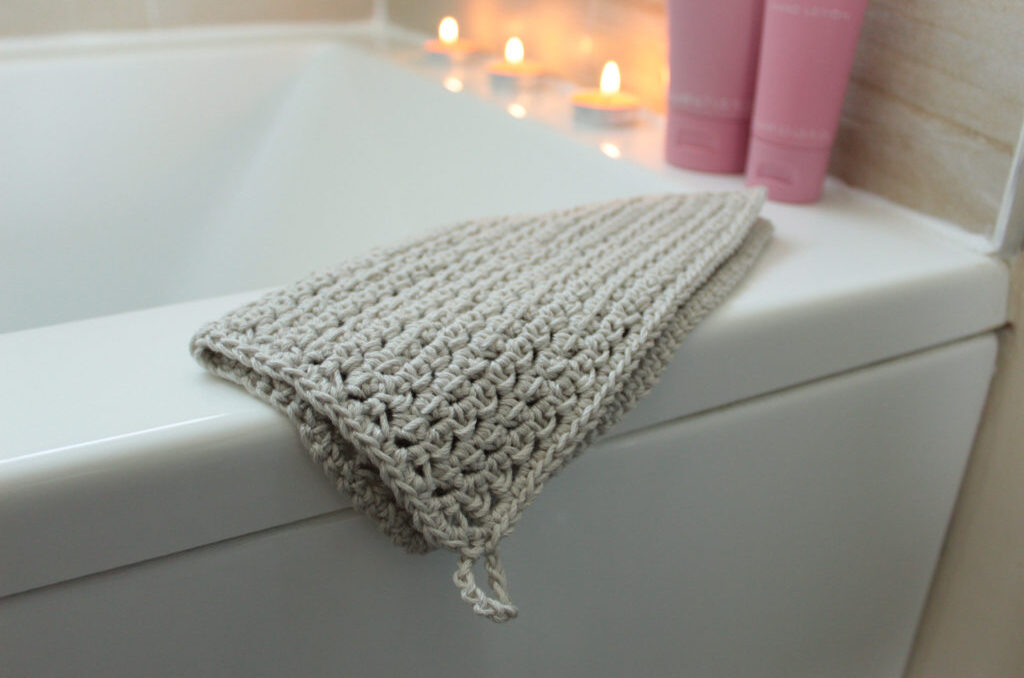 a crochet washcloth with a hanging tab resting on the side of a bathtub with lit tea candles in the background