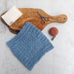 a blue crochet dishcloth on a wooden board with a sponge, scissors and measuring tape