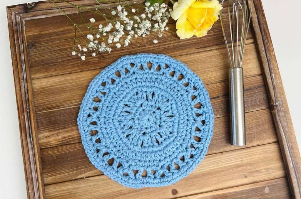 a round crochet dishcloth in blue on a tray with a small whisk