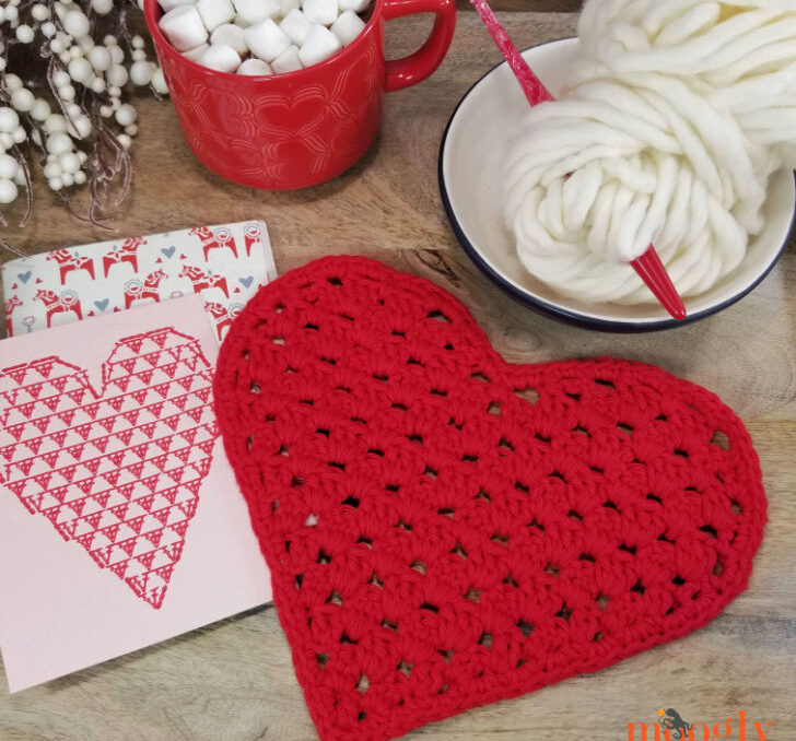 a red crochet heart dishcloth pattern and yarn on a table with a mug.
