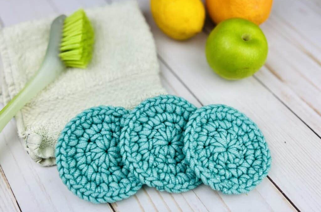 3 blue crochet scrubby rounds on a table with a dish towel and scrub brush