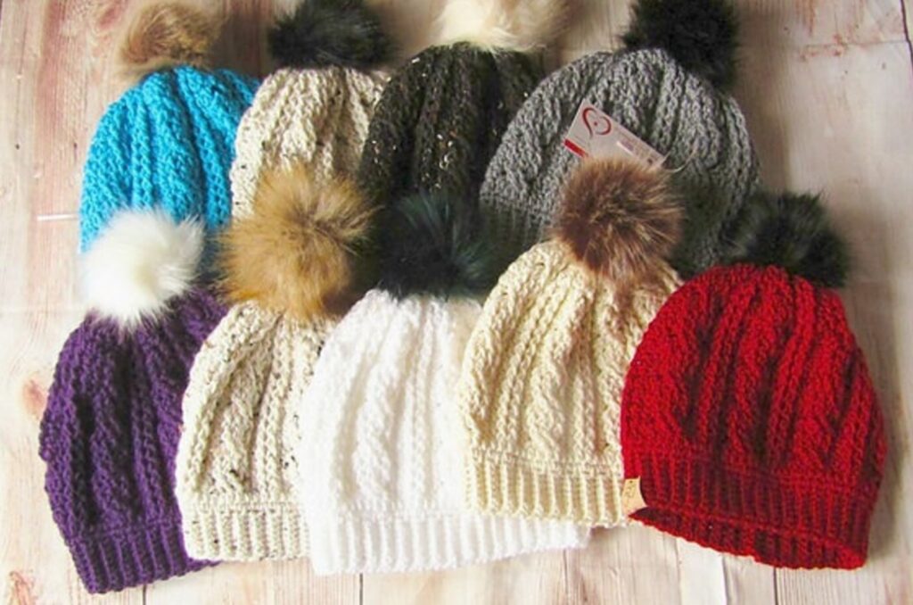 9 crochet cable hats with fur pom poms laid together on a table
