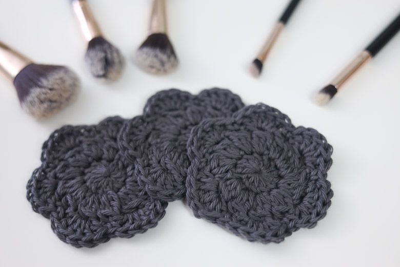 3 crochet face scrubbies shaped like flowers on a table with makeup brushes