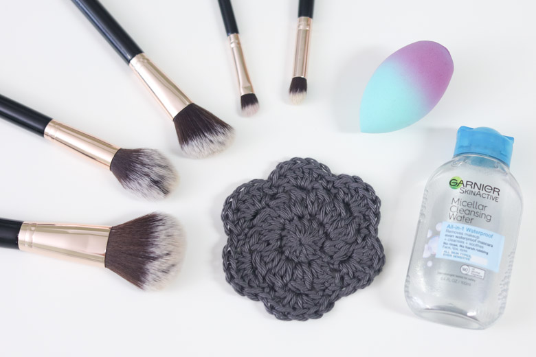 a gray crocheted flower face scrubby pictured with makeup brushes, a makeup sponge, and micellar water