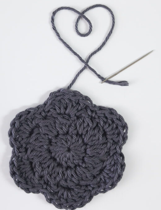 a gray crochet makeup remover pad with the yarn end and needle making a heart