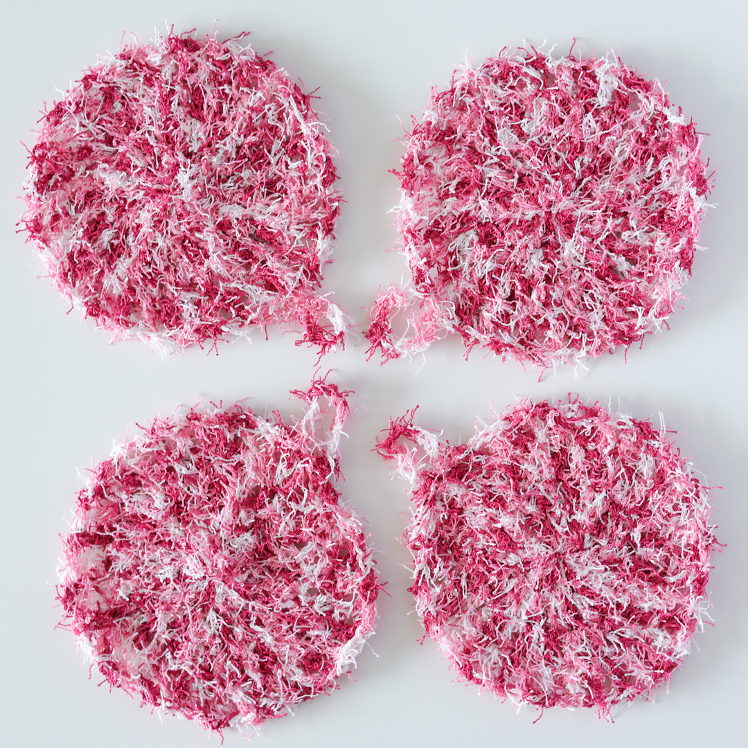 Four pink crochet kitchen scrubbies arranged in a square on a white table