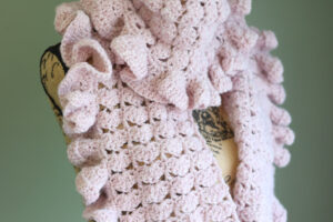 A pink crochet ruffle scarf on a mannequin in front of a green wall.