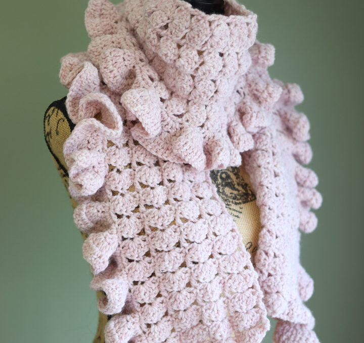 A pink crochet ruffle scarf on a mannequin in front of a green wall.