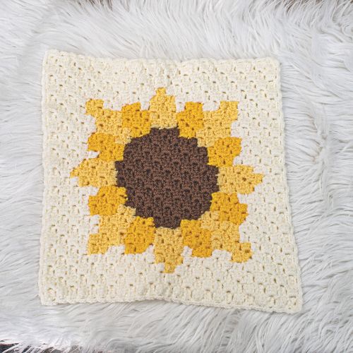 Crochet afghan square that displays a sunflower. Afghan square is on a white textured background. 