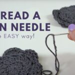 hand holding a threaded yarn needle on a table with gray crochet flowers and text reading How to Thread a Yarn Needle the Easy Way!