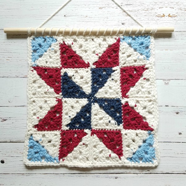 Red, white, navy and light blue crochet granny square wall hanging displayed on white wood background. 