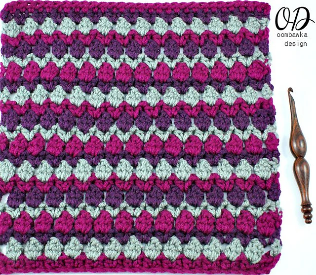A green, purple, and burgundy colored crochet afghan square next to a wooden crochet hook on white background. 