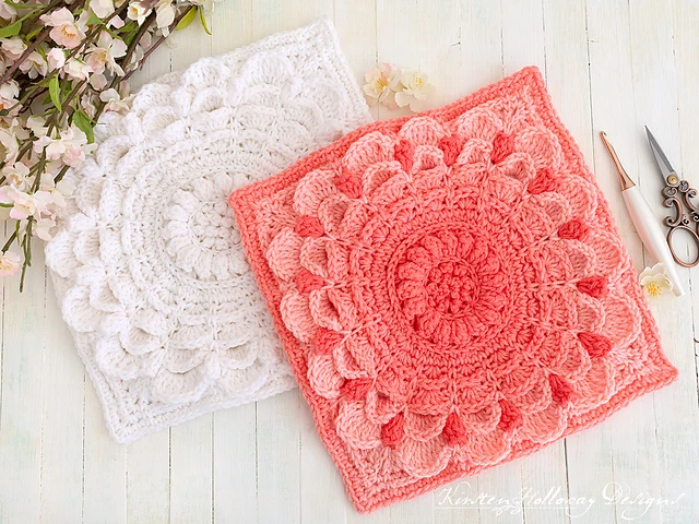 One white and one pink crocheted afghan square that has a textured but floral motif  on a white table.
