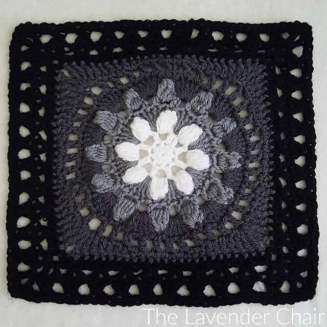 A white, light gray, dark gray, and black crocheted flower motif afghan square on a white background. 