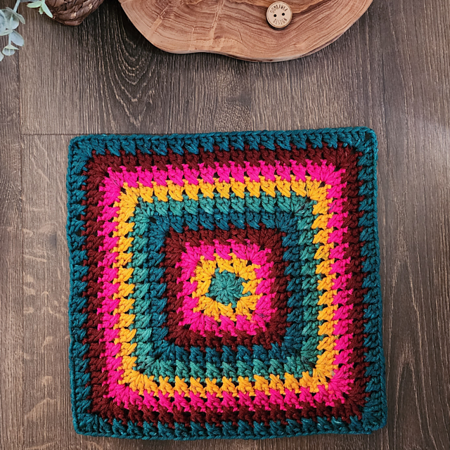 A crocheted blue, yellow, pink, burgundy and blue granny square on a table. 