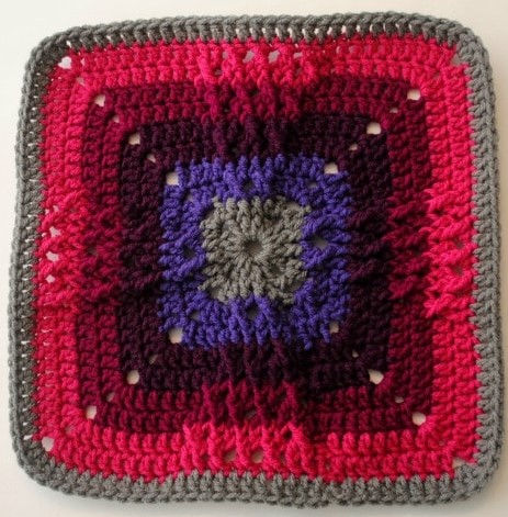 A gray, purple, black, maroon, pink and gray afghan square with textured stitches that mimic a windmill shape. 
